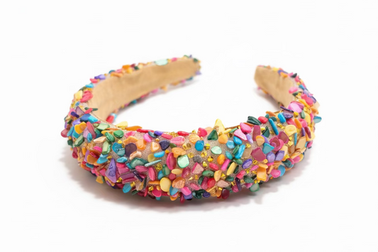 A handmade headband wrapped in brown paper and covered with colorful glitter and sequins, resting on a dark wooden table.