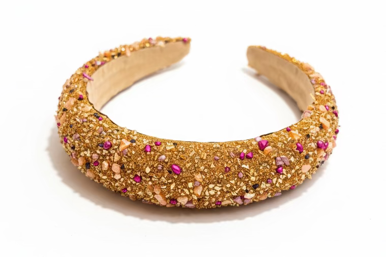A wide, chunky headband covered in textured gold glitter and decorated with scattered pink and metallic rhinestones, resting on a dark wooden surface.