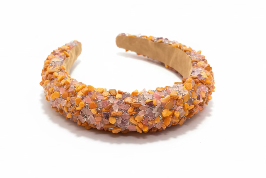A chunky open-front cuff headband covered in textured orange, pink, and gold glitter flakes, resting on a dark wooden surface.